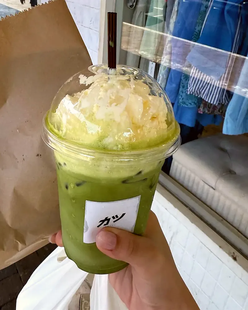 An iced matcha with cream on top, held up outside with a takeaway bag