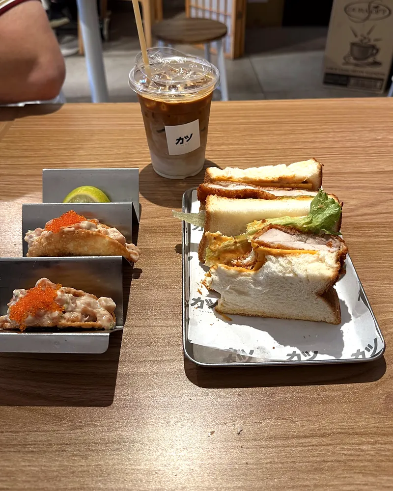 A katsu sando, two sashimi tacos and an iced latte on a table inside the café